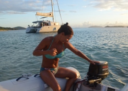 Woman starting a boat motor on the ocean, with a catamaran anchored nearby at sunset. Perfect for powerboating adventures.