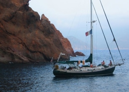 Sailboat anchored near rocky shoreline with two people on board, enjoying the serene coastal view.