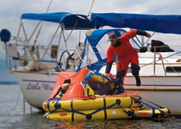 Man in red suit stepping onto a liferaft from a sailboat named Little Wing on calm waters.