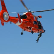 Coast Guard helicopter performing a daring search and rescue operation over open water under clear blue skies.