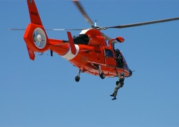 Coast Guard helicopter performing a daring search and rescue operation over open water under clear blue skies.