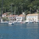 Tranquil marina with moored sailboats in front of colorful seaside village, framed by lush greenery and clear blue waters.