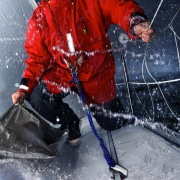 Sailor in red gear harnessed on a wet deck battling waves in stormy weather, showing resilience and skill.