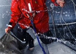 Sailor in red gear harnessed on a wet deck battling waves in stormy weather, showing resilience and skill.