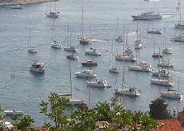 Scenic view of a marina filled with sailboats and powerboats on a sunny day, surrounded by lush green hills and coastal buildings.