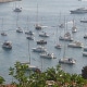 Scenic view of a marina filled with sailboats and powerboats on a sunny day, surrounded by lush green hills and coastal buildings.