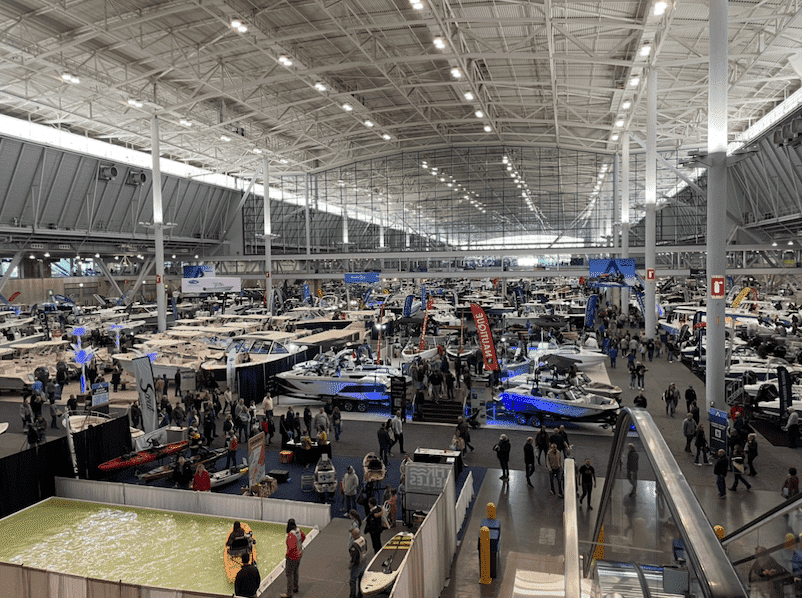 Large indoor boat show with numerous powerboats and attendees exploring the exhibits in a spacious convention center.