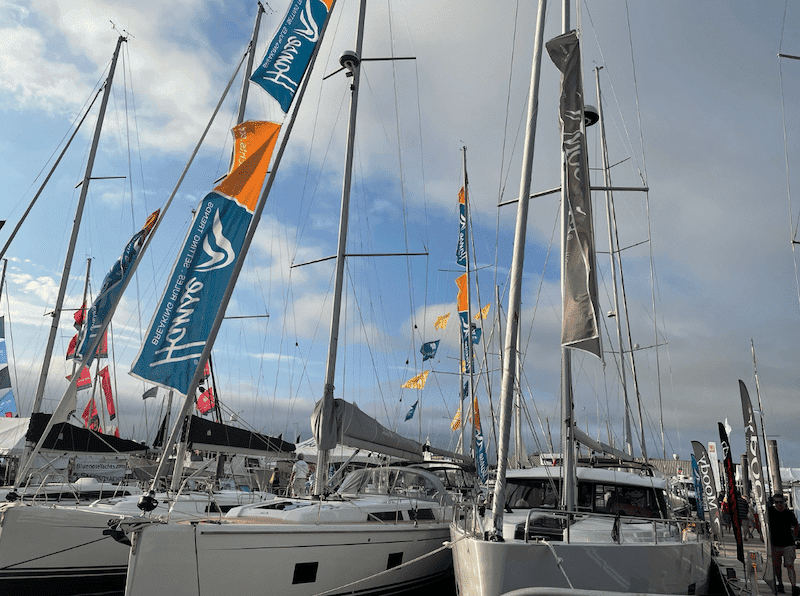 unnamed (46) Sailboats lined up at a marina display vibrant flags under a blue sky, showcasing boating brands and designs.